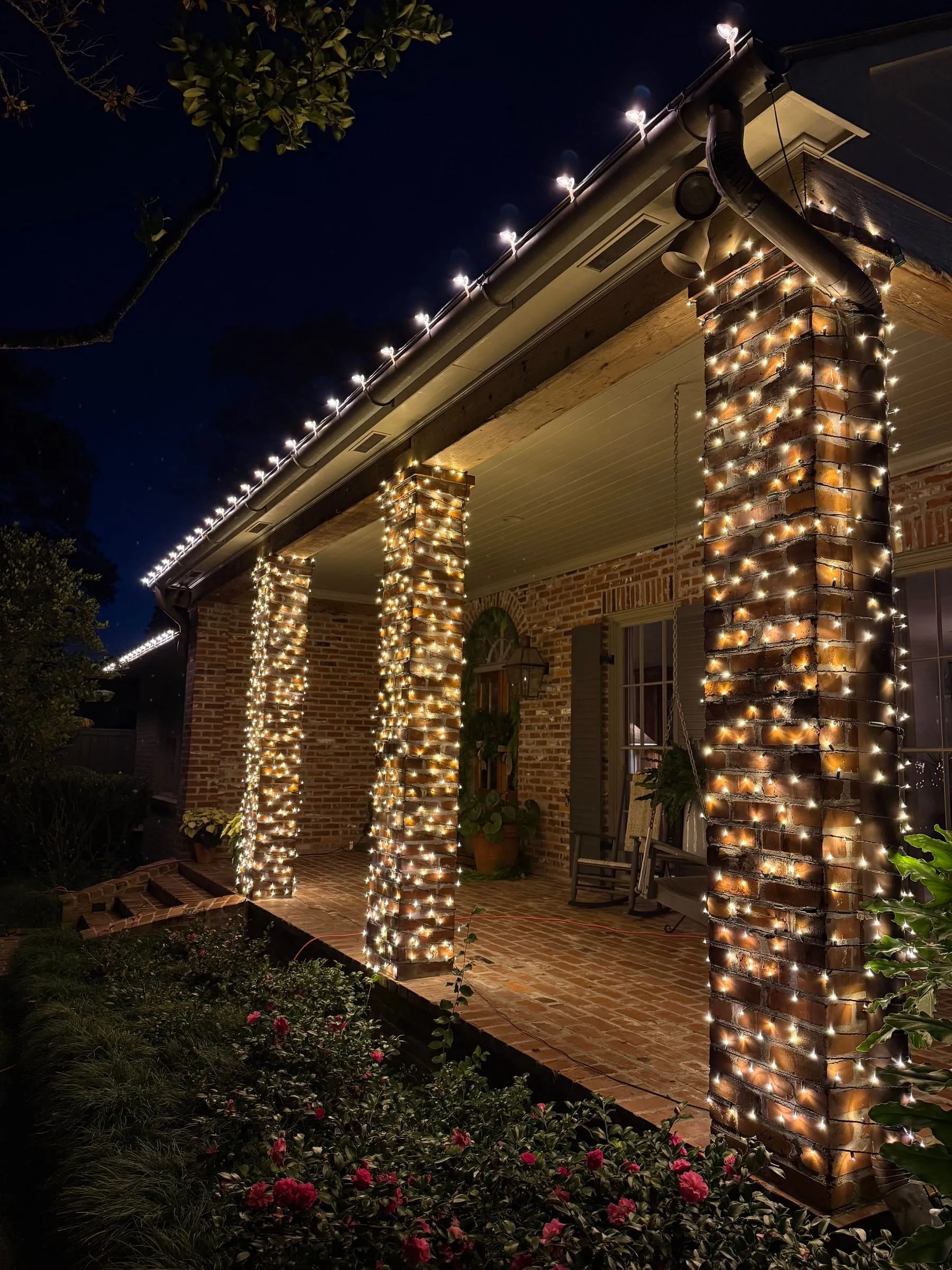 Three brick porch columns wrapped in warm white lights under a lit roofline at night