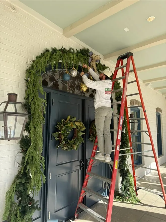 On a ladder hanging garland and ornaments over a front door