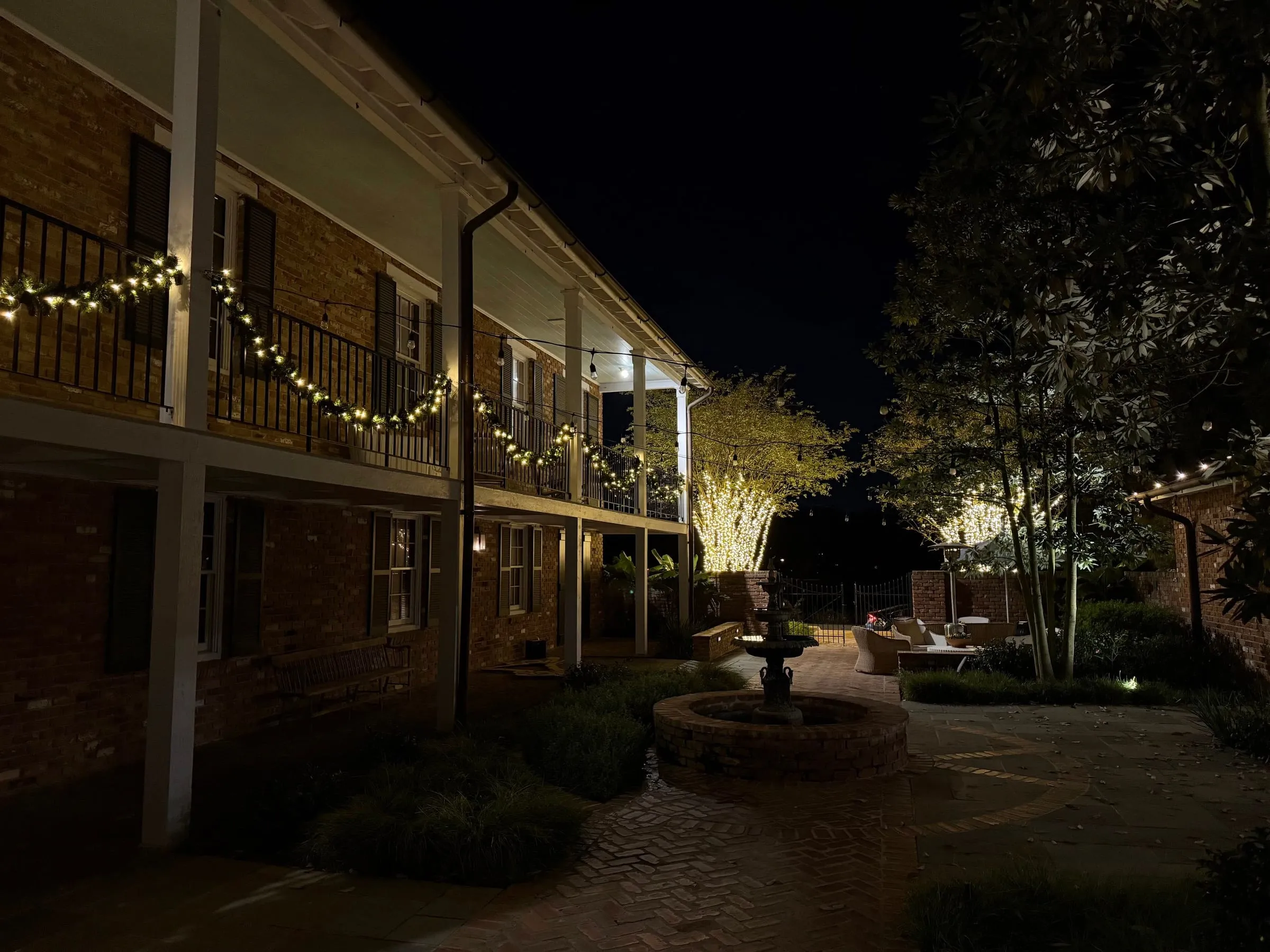 Lit garland along a two-story brick gallery and wrapped trees around a courtyard fountain at night